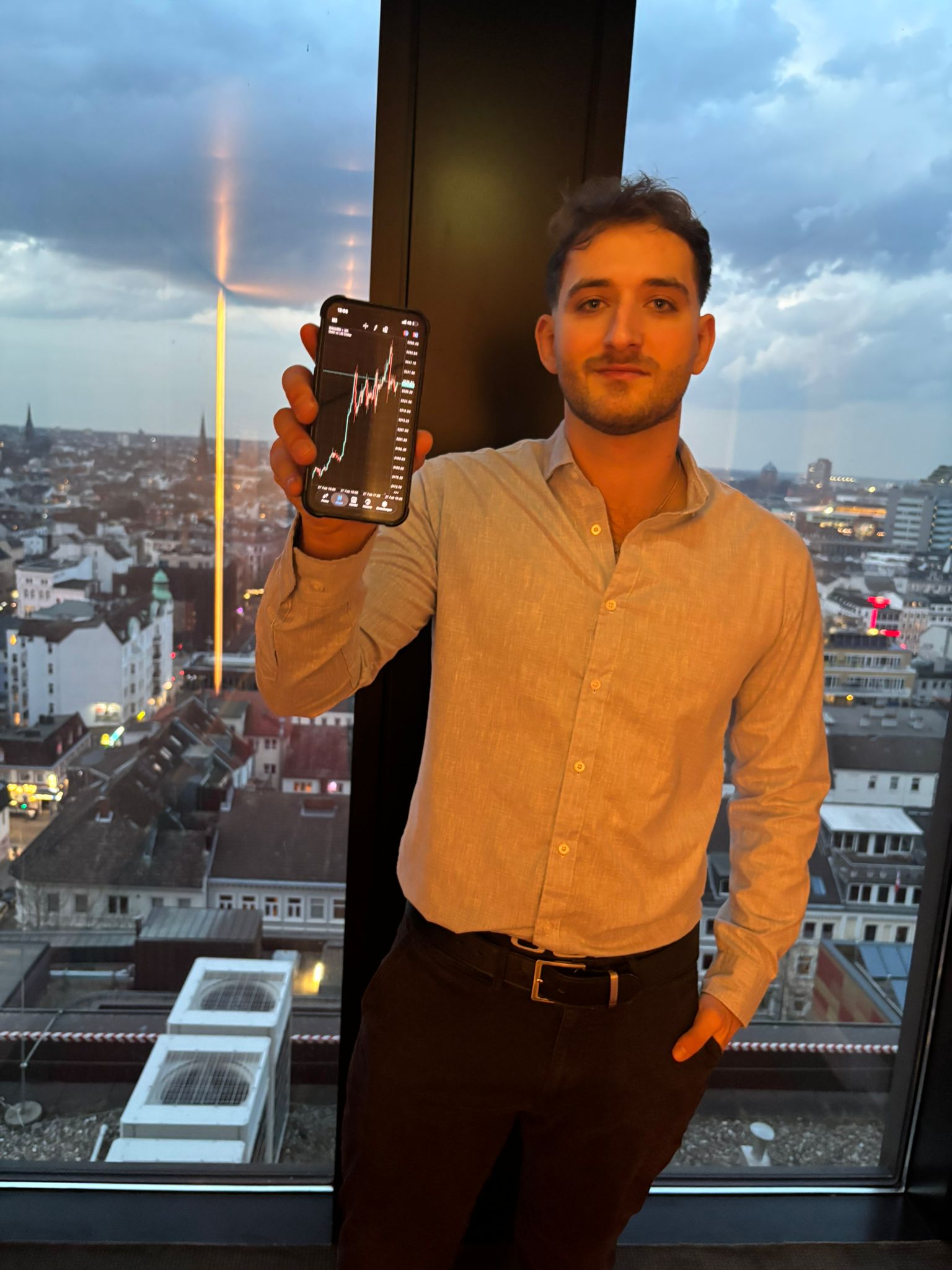 Peer zeigt einen Trading-Chart auf dem Smartphone vor der Stadtskyline bei Dämmerung
